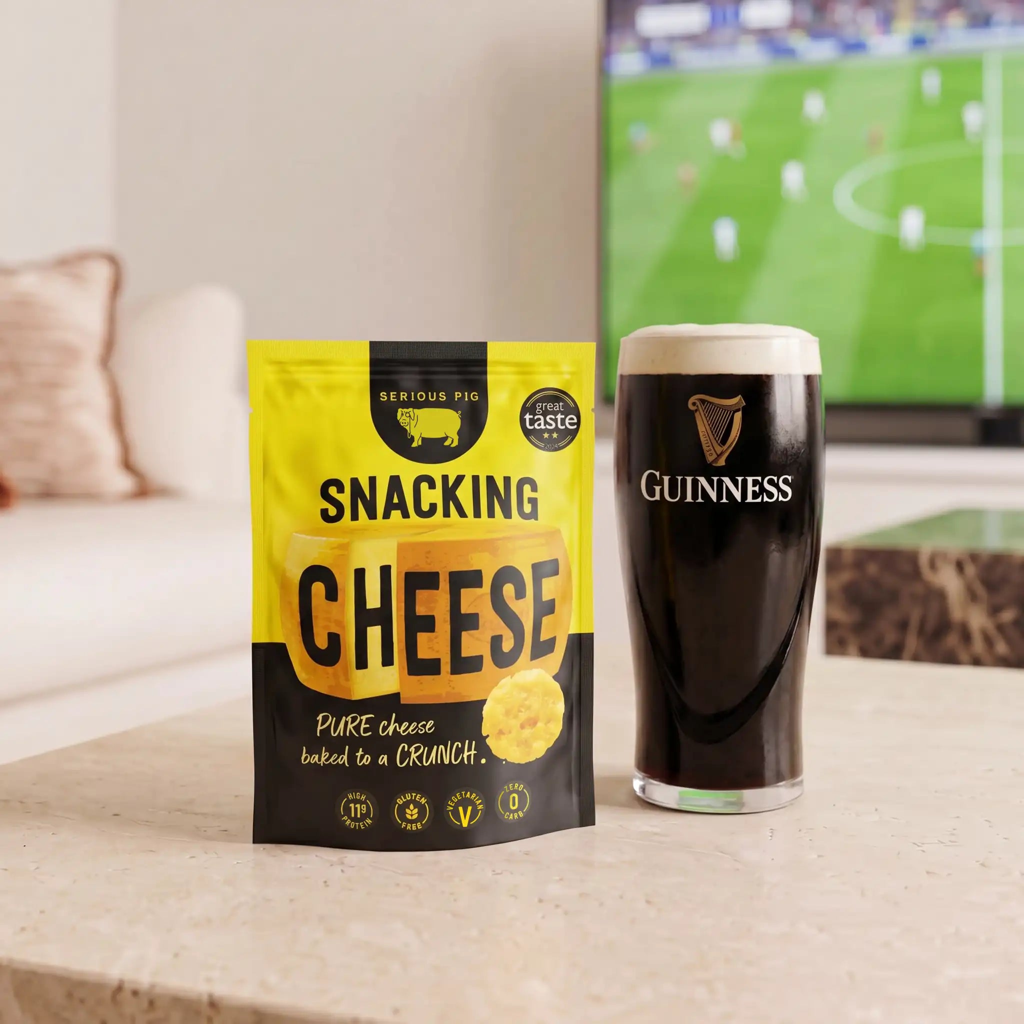 A bag of Snacking Cheese by Serious Pig sits on a light stone countertop next to a pint of Guinness dark beer, with a blurred TV showing a soccer game in the background.
