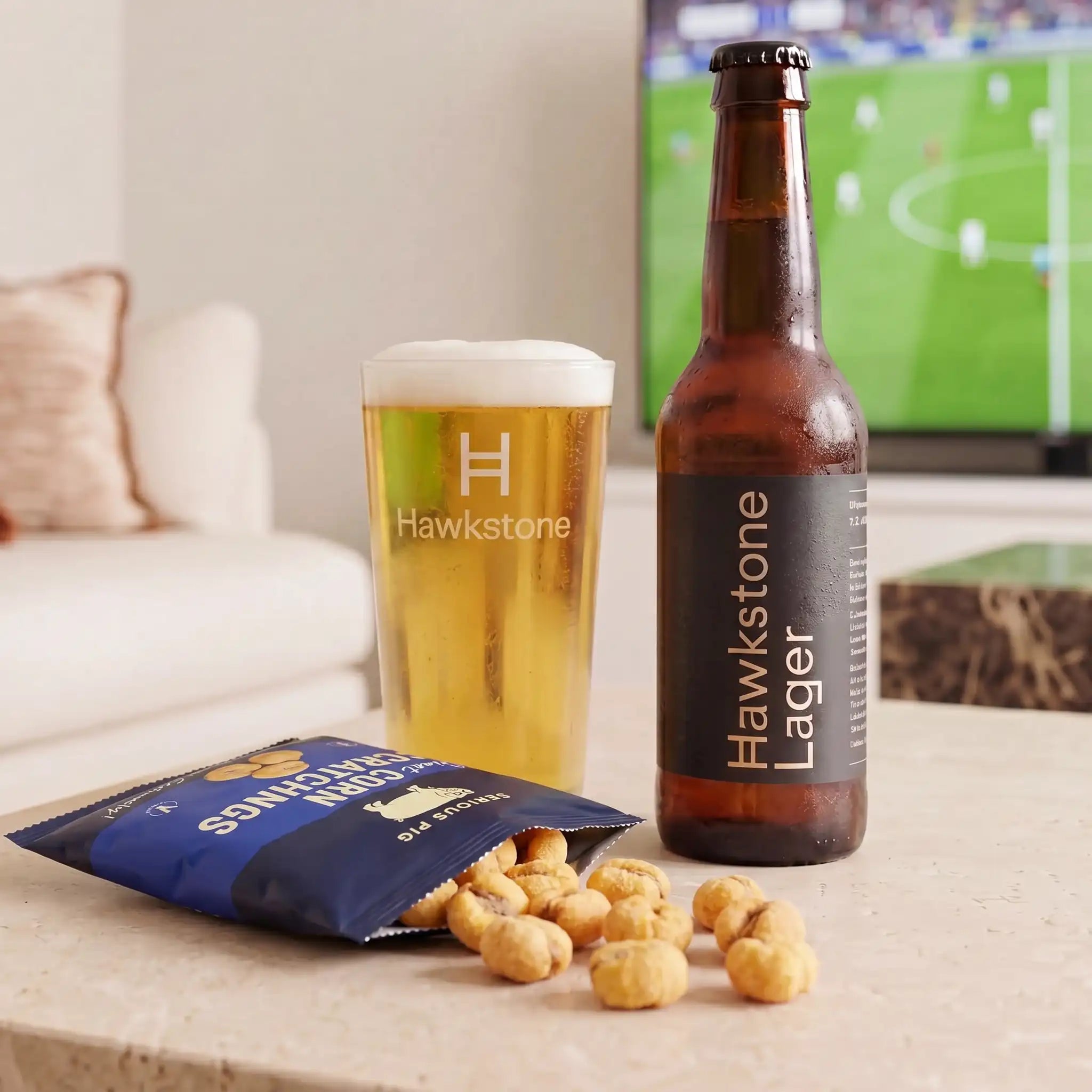 A brown bottle of Hawkstone Lager beer and a glass of golden beer with foam, alongside a blue bag of potato chips with several chips spilled, set on a tabletop in a living room with a blurred TV in the background.
