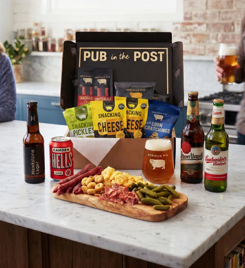 A pub-themed snack and drink gift setup on a kitchen island, featuring a black gift box labeled 'PUB in the POST' with packaged snacks, several beer bottles, a can of Camden Hells beer, a glass of amber beer, and a charcuterie board with crackers, cheese, sausage, beans, and pickles.