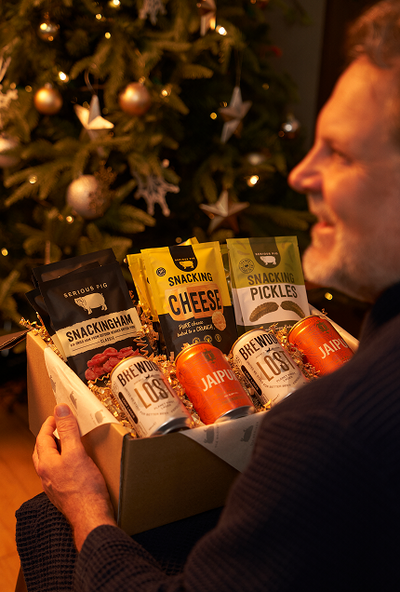 A man holds a gift box filled with snacks and drinks in front of a decorated Christmas tree. The box contains bags of Snacknag Cheese, Pickles, and Spaghetti, along with cans of Rhinowolf Los Angeles and Jaipur.