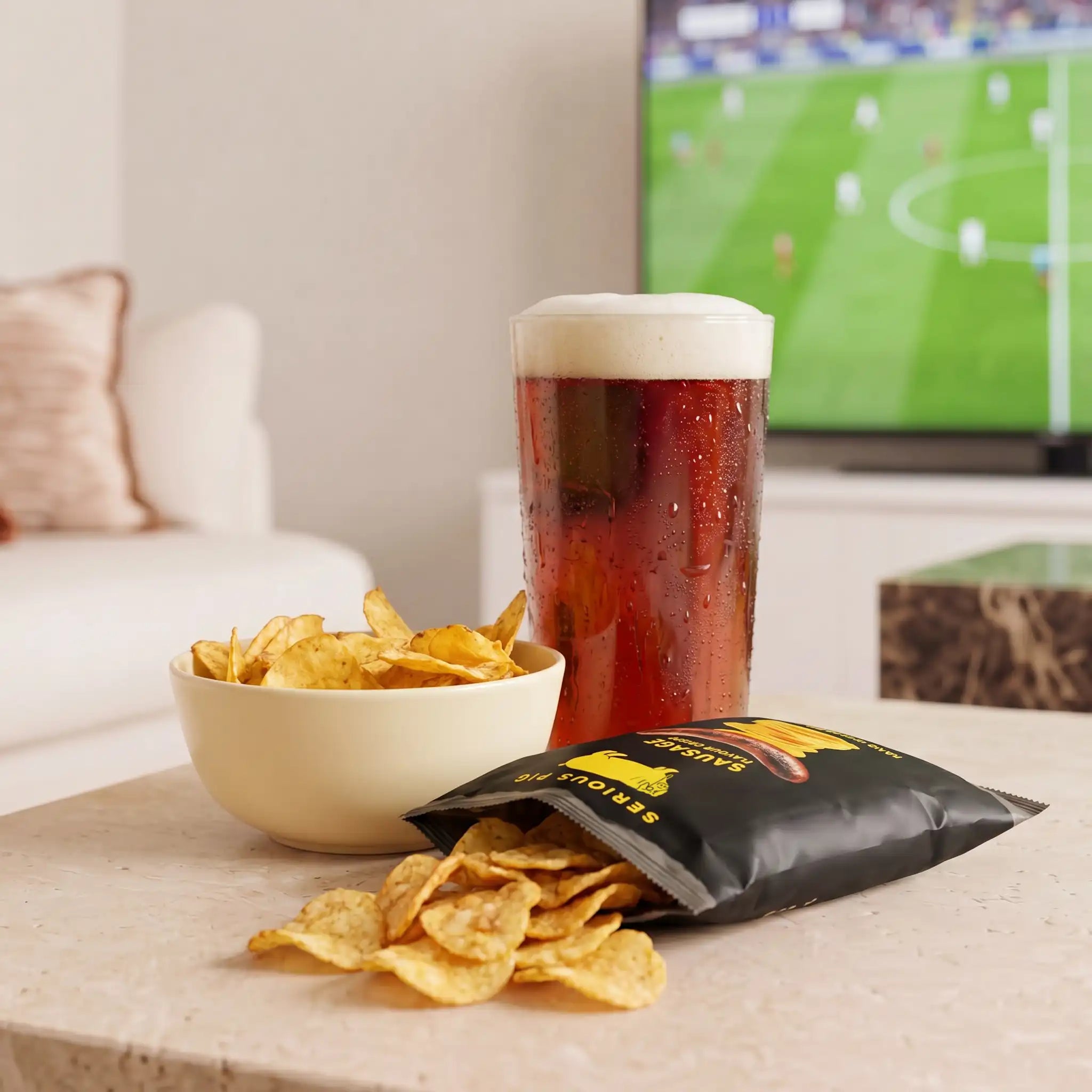 A lifestyle scene showing a bag of potato chips, a bowl of chips, and a glass of beer on a table in a living room, with a TV showing a soccer game in the background.