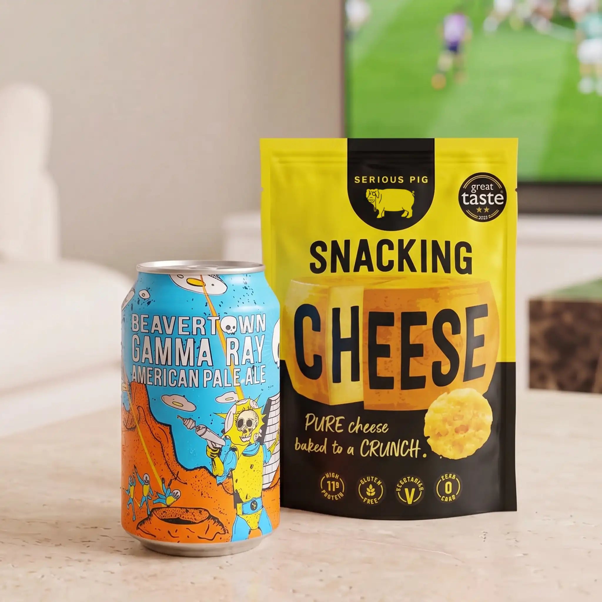 A can of Beavertown Gamma Ray American Pale Ale sits next to a bag of Serious Pig Snacking Cheese on a light beige countertop in a kitchen setting.