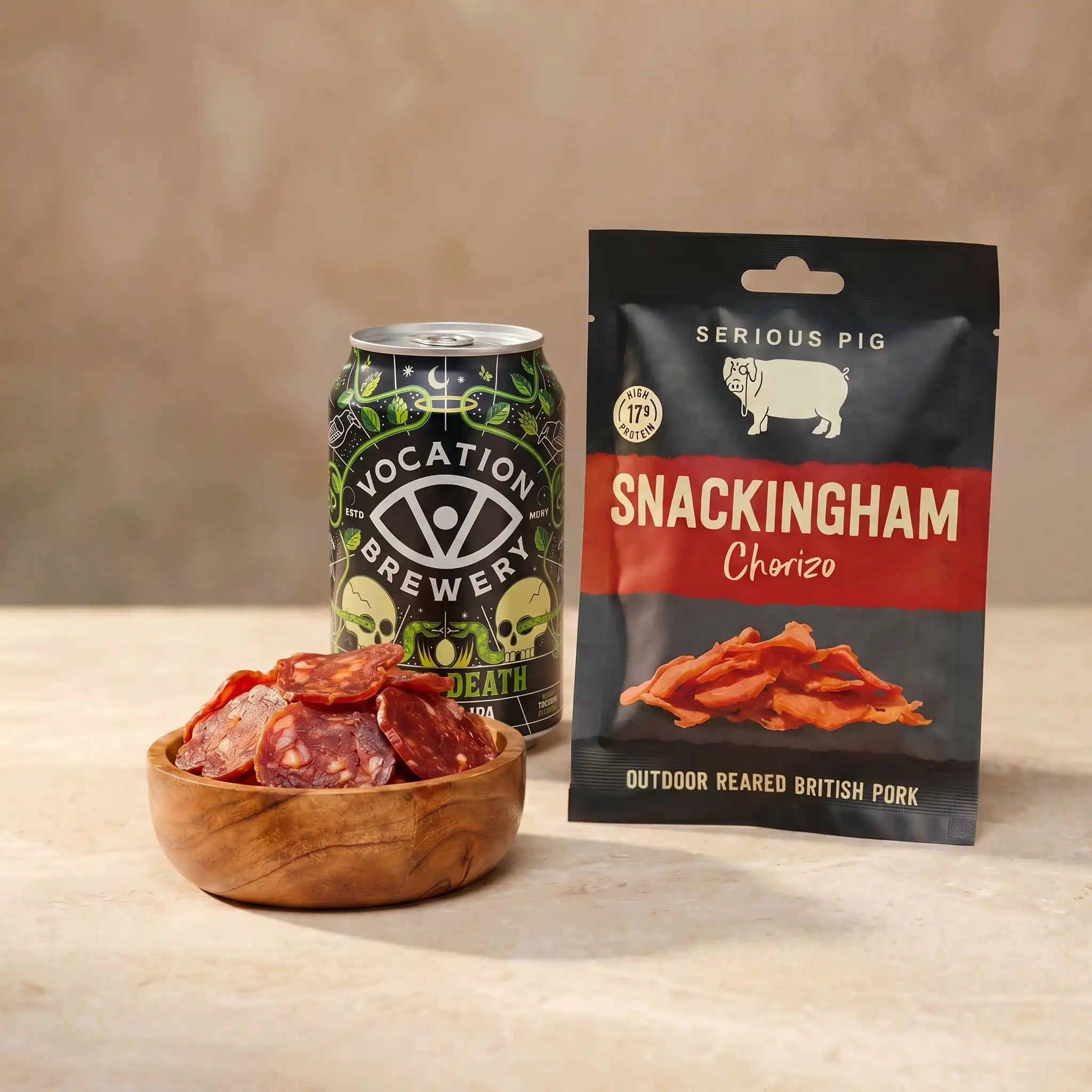 A can of Vocation Brewery Life & Death Ale stands next to a bag of Serious Pig Snackingham Chorizo pork jerky and a small wooden bowl filled with sliced chorizo, set on a beige surface against a neutral beige background.