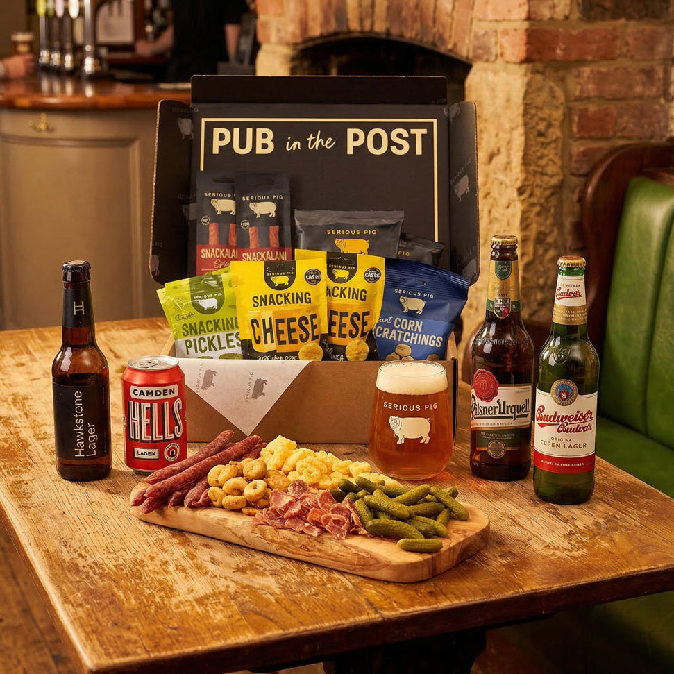 A pub-themed snack and beer gift set displayed on a wooden table in a restaurant setting. The set includes a black gift box labeled 'PUB in the POST' with assorted branded snacks, a can of Hells Hell's, a bottle of Sam Adams London Dry, a bottle of Budweiser, and a beer glass, alongside a charcuterie board with green beans, cheese, sausage, and crackers.