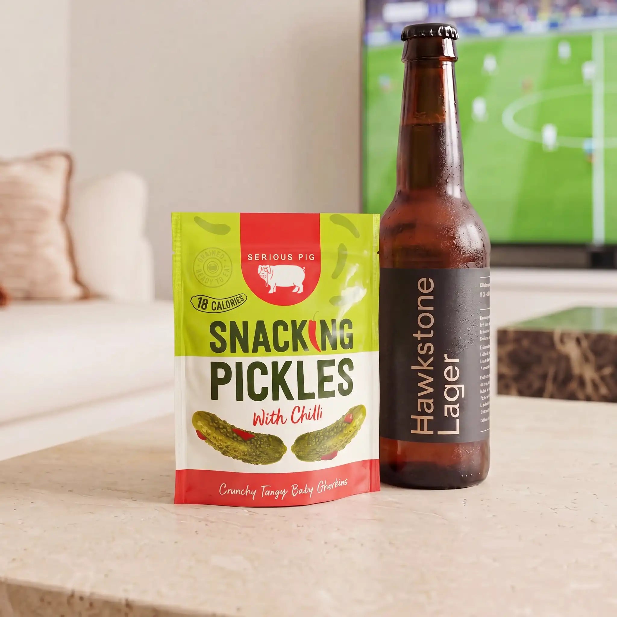 A bag of Serious Pig Snacking Pickles with Chili stands next to a brown Hawkstone Lager beer bottle on a light stone countertop in a living room setting, with a blurred TV in the background.